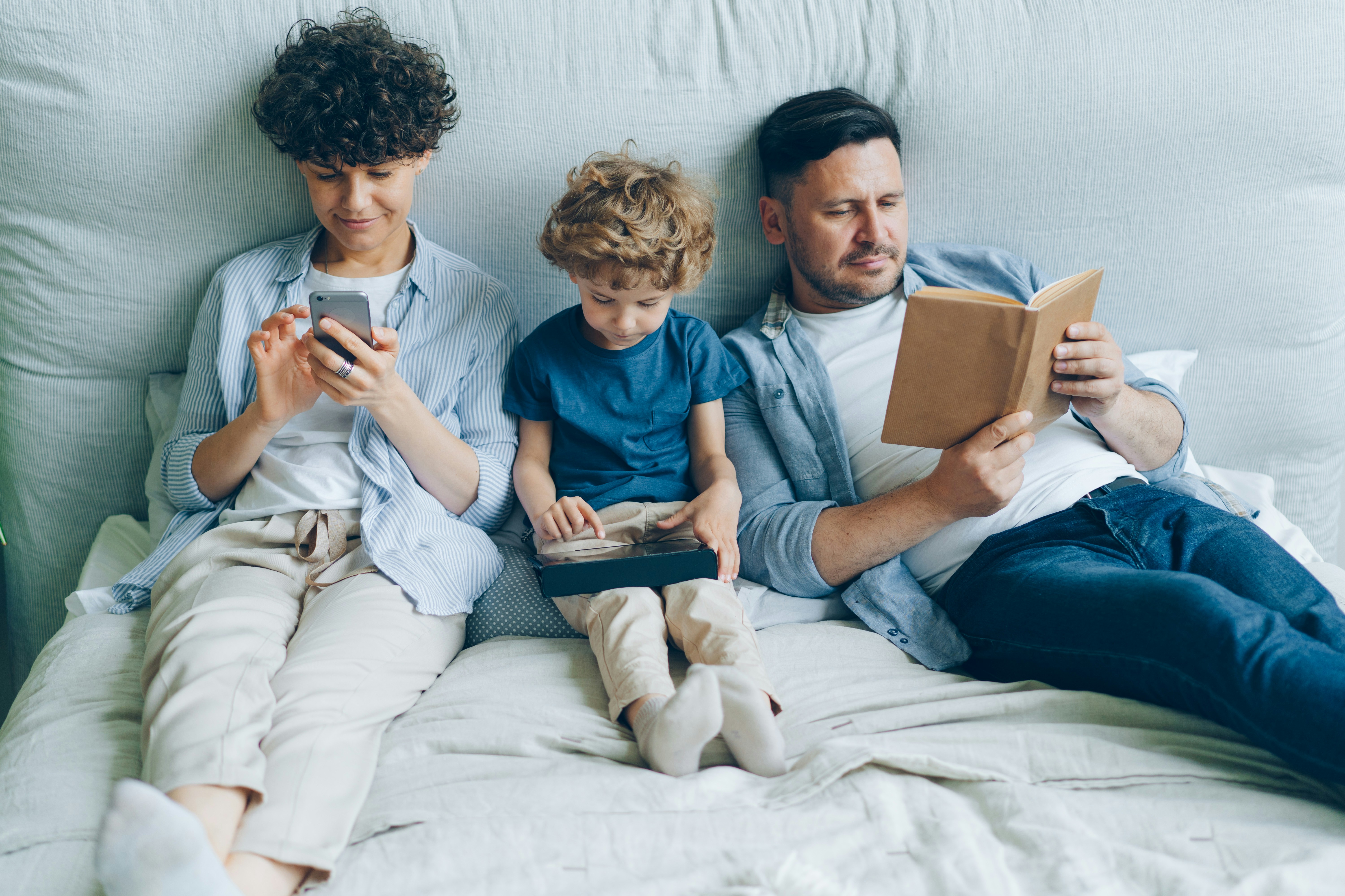 Family relaxing together — mom on phone, child on tablet, dad reading a book